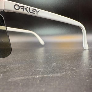 Może przedstawiać: Białe okulary przeciwsłoneczne Oakley z ciemnymi soczewkami. Logo Oakley jest nadrukowane na białej oprawce. Okulary przeciwsłoneczne mają zakrzywioną konstrukcję i są na ciemnym tle.