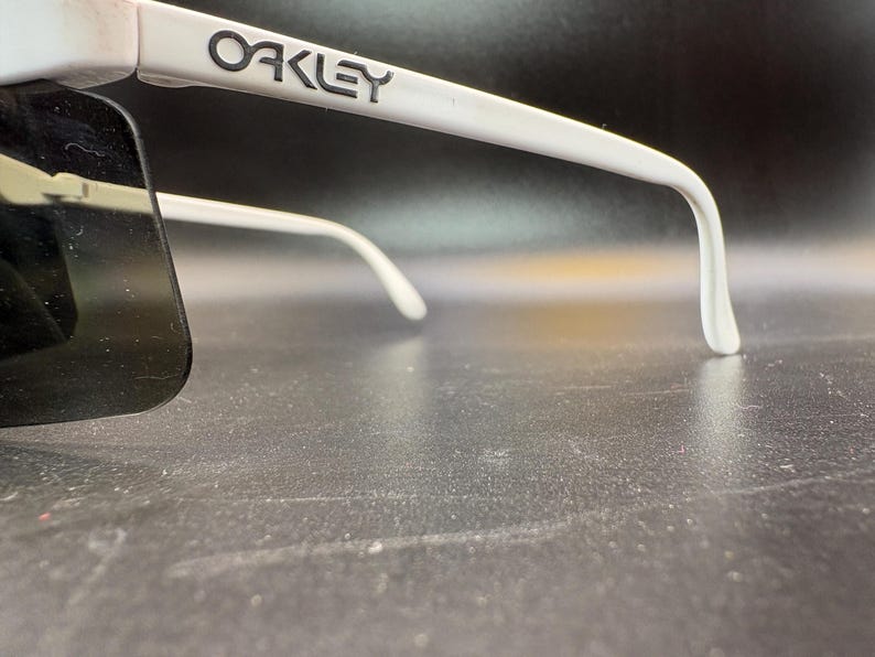 Może przedstawiać: Białe okulary przeciwsłoneczne Oakley z ciemnymi soczewkami. Logo Oakley jest nadrukowane na białej oprawce. Okulary przeciwsłoneczne mają zakrzywioną konstrukcję i są na ciemnym tle.