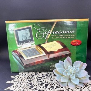 May include: A rectangular product box for an Expressive World Time Clock and Calculator Desk Set. The box features a dark wood grain finish, a digital clock display, a calculator keypad, a pen holder, and a yellow sticky note pad.