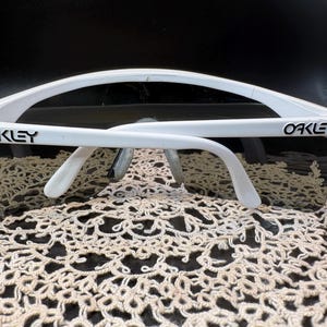 Może przedstawiać: Białe okulary przeciwsłoneczne Oakley z ciemnymi soczewkami. Logo Oakley jest widoczne na białej oprawce. Okulary przeciwsłoneczne leżą na kremowej, ażurowej powierzchni.