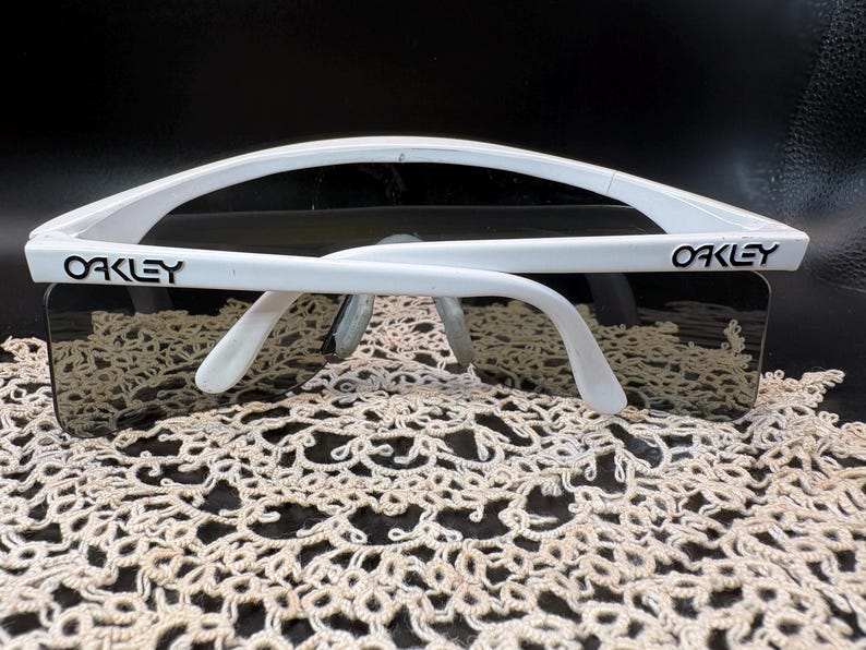Może przedstawiać: Białe okulary przeciwsłoneczne Oakley z ciemnymi soczewkami. Logo Oakley jest widoczne na białej oprawce. Okulary przeciwsłoneczne leżą na kremowej, ażurowej powierzchni.