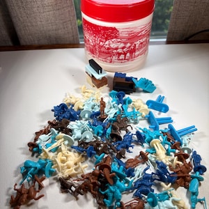 May include: A white plastic storage container with a red lid and a red landscape illustration, accompanied by a large collection of small, multi-colored plastic toy figures including horses, riders, and various building block components.