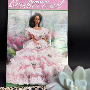 May include: Annie's Crochet To Go! magazine, April/May 2000, number 122. The cover features a doll wearing a tiered, ruffled, light pink and white crocheted gown adorned with small fabric rosebuds, set against a garden background.