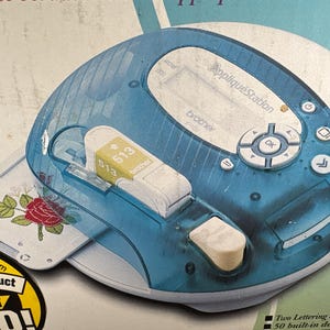 May include: A blue and white Brother AppliquéStation machine with a white and yellow 513 cartridge. The machine has a clear blue top and a white base with a floral design. The machine has buttons and a screen.