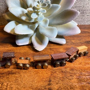 May include: A miniature wooden toy train consisting of a locomotive and four connected cars. Each car features a dark brown wooden body with small, metallic gold-colored wheels. The train is arranged in a line, showcasing the detailed wood grain texture on the roofs and sides of the individual carriages.
