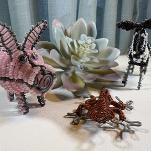 May include: A collection of wire and bead animal sculptures. The sculptures include a pink beaded pig, a black and white beaded zebra, and a brown wire frog. A faux succulent plant is in the background.