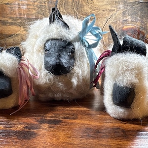 May include: Three handcrafted sheep figurines with fluffy white wool bodies and black faces and ears. Each sheep has a decorative ribbon bow in shades of pink and blue. The figurines are displayed on a wooden surface.