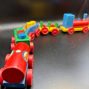 May include: A wooden toy train set with multiple cars. The engine is red and blue, with a yellow smokestack. The cars are painted in bright colors: red, green, yellow, and blue. The first car has the Esso logo. The train has red wheels.