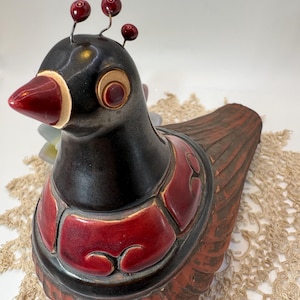 May include: A ceramic bird figurine featuring a black head and neck with three small red bead-like antennae. The body is decorated with glossy red panels over a textured, dark reddish-brown base. The bird has a red beak and a circular eye detail.