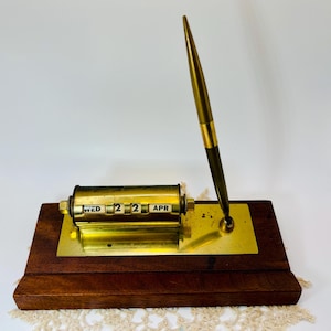 May include: A vintage desk calendar with a pen holder. The calendar displays "WED 22 APR" in black text on a gold-colored cylindrical dial. The pen is gold and black, and the base is wood and gold-colored metal.