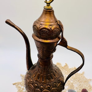 May include: A copper-colored, ornate pitcher with a long spout and handle. The pitcher features detailed embossed patterns and a decorative top with a crescent moon and star design. The pitcher is likely used for serving beverages.