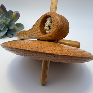 May include: A wooden spinning top with a rounded base and a handle. The top has a carved design with a small opening containing a white string. The wood has a natural, light brown color.