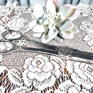 May include: Silver-toned vintage scissors with ornate handles and a decorative floral center. The scissors are resting on a white lace tablecloth, with a white and pink succulent in the background. The scissors are approximately 8 inches long.