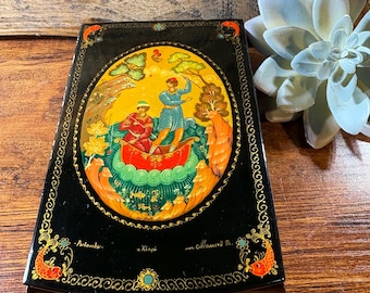 May include: A black lacquered box with a vibrant oval painting depicting figures in a boat, surrounded by trees and a yellow sky. The box has gold trim and decorative fish accents. The text on the box is in Russian.