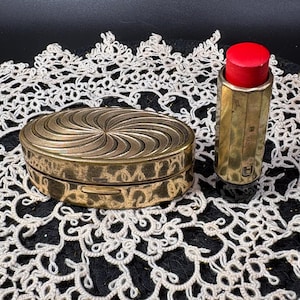 May include: An antique gold-toned compact with a spiral design on the lid and a matching lipstick case with a red lipstick. The items are displayed on a white lace doily against a black background.