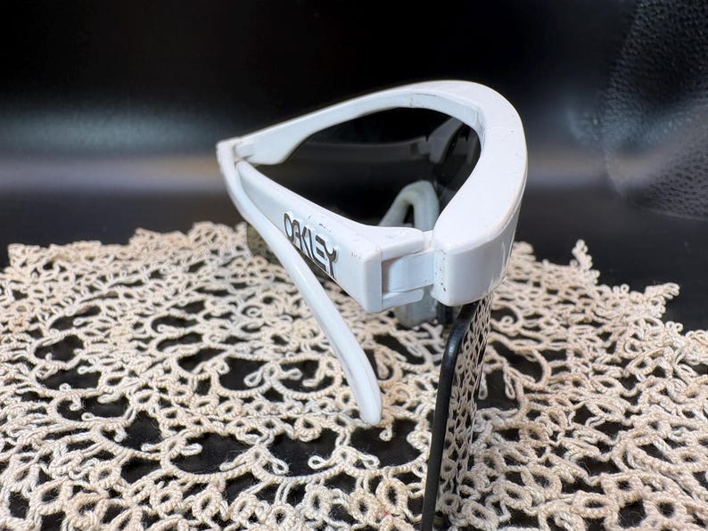 Może przedstawiać: Białe okulary przeciwsłoneczne Oakley z ciemnymi soczewkami. Logo Oakley jest widoczne na oprawce. Okulary przeciwsłoneczne są umieszczone na ciemnym tle i kremowej koronkowej serwetce.