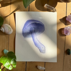May include: A watercolor painting of a blue mushroom on a white paper. The artwork is surrounded by natural elements, including a seashell, crystals, and greenery, all set against a wooden background. The mushroom has a detailed cap and stem.