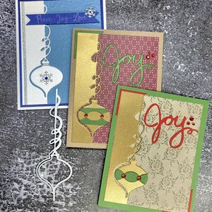 May include: Handmade Christmas cards with ornament cutouts. One card has the words "Peace Joy Love" on a blue banner. Other cards feature the word "Joy" in cursive. The cards have gold, green, red, and blue accents.
