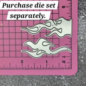 May include: Two silver metal die cuts in the shape of flames. The die cuts are on a pink cutting mat with a ruler in inches. The text "Purchase die set separately." is at the top of the image.