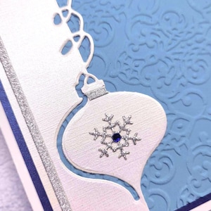 May include: A white card with a blue background and a silver ornament cut out. The ornament has a snowflake with a blue jewel in the center. The card has the text "Peace, Joy, Love" written on it.