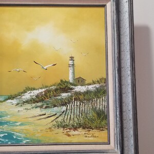 May include: A framed oil painting of a lighthouse on a sandy beach with a yellow sky and seagulls flying overhead. The painting is signed "Ken Fisher".