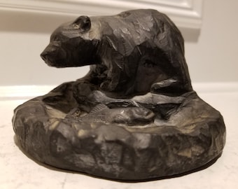 May include: A black carved stone bear figurine sitting on a stone base with a carved out area in the center. The bear is looking to the left.