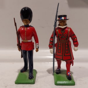May include: Two toy soldier figurines, one in a red uniform with a black hat and a rifle, the other in a red and gold uniform with a black hat and a spear.