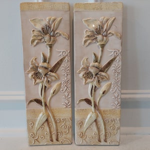 May include: Two rectangular ceramic wall plaques with raised floral designs. Each plaque features a light pink background with a brown and white floral design. The flowers are stylized and have a 3D effect.