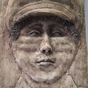 May include: A beige plaster relief sculpture of a person's face. The person is wearing a cap with a textured design. The sculpture has a textured surface and a neutral expression.