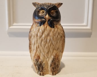 May include: A ceramic owl figurine with brown and black markings. The owl has large, round eyes with yellow pupils. The owl is standing on a white surface.