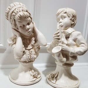 May include: A pair of white ceramic figurines, one depicting a girl covering her ears and the other a boy playing a saxophone. Both figurines are on pedestal bases.