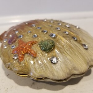 May include: A white seashell trinket box with a gold iridescent finish. The box is decorated with rhinestones, a starfish, and a green seashell.