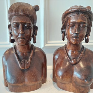 May include: Two wooden sculptures of female busts. The sculptures are carved from dark brown wood and feature detailed facial features, earrings, and necklaces. The busts have a smooth, polished finish and are displayed side by side.