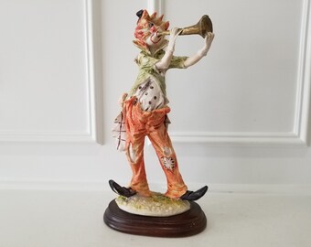 May include: A whimsical figurine of a clown playing a trumpet. The clown wears a green jacket, orange pants with patches, and a black hat. The figurine stands on a wooden base with a grassy area.