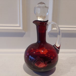 May include: A ruby red glass decanter with a clear handle and stopper. The decanter has a round base and a long neck with a flared lip. The stopper is faceted. Etched floral and architectural designs adorn the decanter.