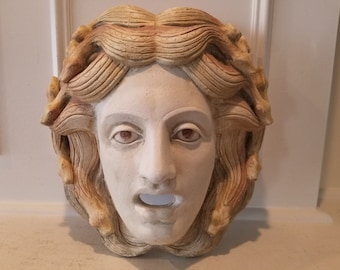 May include: A white plaster mask of a woman's face with brown hair sculpted in a wavy style. The mask has a wide-open mouth and red painted eyes.