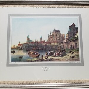 May include: A vintage print of Cologne, Germany, featuring a detailed depiction of the city's skyline, including the Cologne Cathedral, with a river in the foreground and boats on the water. The print is framed in a silver frame with a lace border.