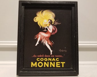 May include: A framed vintage advertisement for Cognac Monnet featuring a woman in a red dress holding a glass of cognac. The text reads "...du soleil dans un verre... COGNAC MONNET".