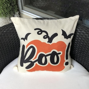 May include: A square off-white throw pillow featuring a central orange pumpkin graphic with a black stem. The word Boo! is printed in large black script across the pumpkin, accompanied by two small black bat silhouettes on either side.