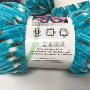 May include: A skein of bulky weight 5 chenille yarn in a variegated pattern of turquoise, dark green, and white. The white paper label features care instructions, a 10 cm by 10 cm gauge swatch guide, and the text Machine Wash Warm, Dry flat.