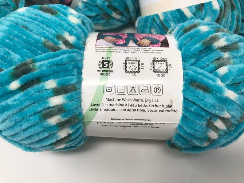 May include: A skein of bulky weight 5 chenille yarn in a variegated pattern of turquoise, dark green, and white. The white paper label features care instructions, a 10 cm by 10 cm gauge swatch guide, and the text Machine Wash Warm, Dry flat.