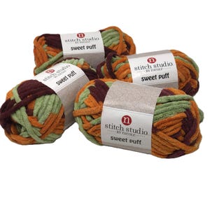 May include: Four skeins of Stitch Studio by Nicole Sweet Puff yarn, each featuring a variegated blend of soft sage green, burnt orange, and deep maroon fibers. Each skein is wrapped in a white paper label with the brand name and product text.