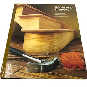 May include: A book titled "Floors and Stairways" featuring a wooden staircase, a hammer, and a small orange brush. The book's cover has text about flooring and stair repair. The hammer is silver and black.