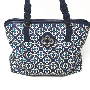 May include: A navy blue tote bag with a geometric teal and white pattern. The bag has navy blue braided handles and trim. A silver-colored emblem is centered on the front. The text "KNOT SIMPLE Designs" is in the upper left corner.
