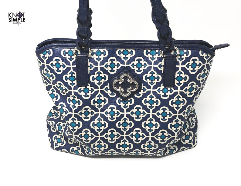 May include: A navy blue tote bag with a geometric teal and white pattern. The bag has navy blue braided handles and trim. A silver-colored emblem is centered on the front. The text "KNOT SIMPLE Designs" is in the upper left corner.