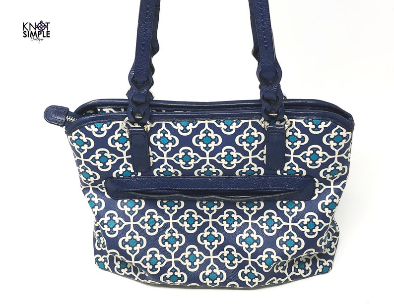May include: A navy blue tote bag with a white and teal geometric pattern. The bag has navy blue handles and trim, with a zippered pocket on the front. The bag has the text "Knot Simple Boutique" in the upper left corner.