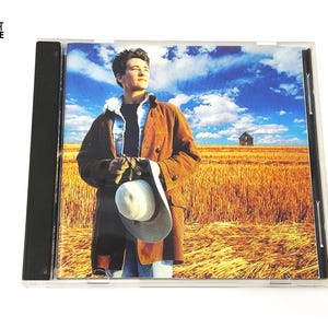 May include: CD case featuring a man in a brown coat and a white hat, set against a backdrop of a wheat field under a blue sky with clouds. The CD case is in a clear plastic jewel case.