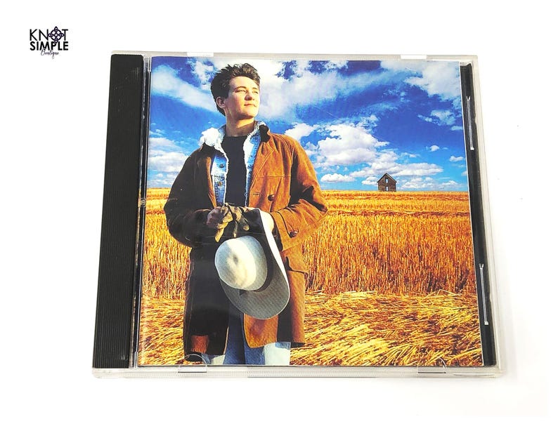 May include: CD case featuring a man in a brown coat and a white hat, set against a backdrop of a wheat field under a blue sky with clouds. The CD case is in a clear plastic jewel case.