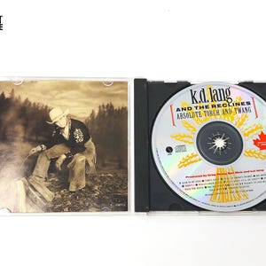 May include: A K.D. Lang and the Reclines "Absolute Torch and Twang" CD in a black jewel case. The CD has a silver surface with the album title and a Canadian maple leaf. The album art features a sepia-toned image of a person in a cowboy hat.
