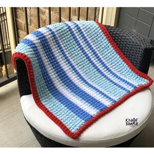 May include: A handmade crochet blanket featuring a textured bobble stitch pattern with vertical stripes in shades of light blue, medium blue, and white. The blanket is finished with a solid red border and includes the text Knot Simple Boutique.